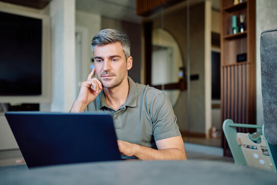 Middle-aged man sitting in his living room, working remotely on a laptop, concentrating and thinking deeply - Powered by Adobe