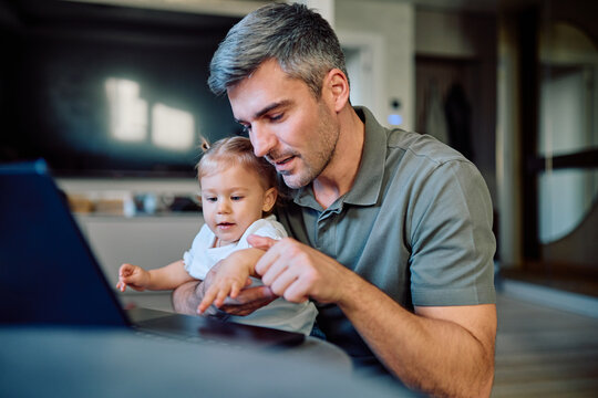 Father holding his baby daughter, showing her content on a laptop screen and pointing with their fingers
