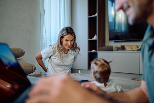 Happy family spending time together, mother smiling and playing with toddler while father work on the laptop