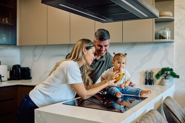 Happy family together in a contemporary kitchen, parents feeding their cute toddler a banana, promoting healthy eating and bonding