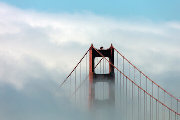 Dense coastal fog drifts across a sweeping suspension bridge, partially veiling its towers in soft morning light.