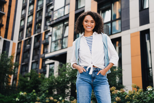 Young woman student stands in front of modern city apartments smiling confidently in casual urban attire conveying lifestyle and travel vibe for stock photography