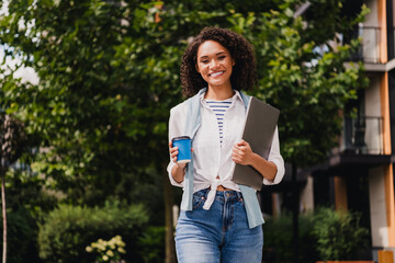 Casual student woman walking outdoors with coffee and laptop smiling in modern city park enjoying work and study on the go