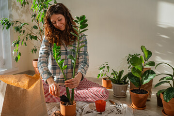 Woman sitting on the floor transplanting a zamioculcas zamiifolia plant into a larger pot, surrounded by several other green houseplants, enjoying her indoor gardening hobby