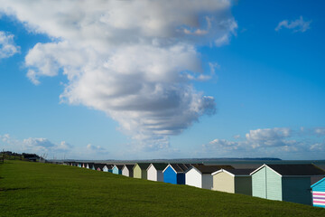 Wooden beach huts painted in different colours stretch into the distance on Tankerton Beach, Whitstable. A large white cloud dominates the blue sky. 