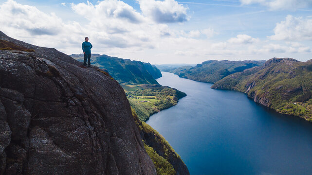 Person standing on cliff above scenic Norwegian fjord, dramatic mountain landscape with deep blue water and cloudy sky, aerial drone view of Ørsdalsvatnet - Powered by Adobe