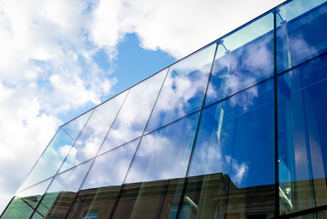 A fragment of a glass-clad building rising toward the sky, with reflections of clouds and an opposite building on its mirrored surface. The image highlights modern architecture, symmetry.