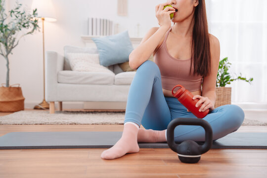 Fit woman resting after home workout, sitting on yoga mat with kettlebell, water bottle, and green apple healthy lifestyle, self-care, and recovery.