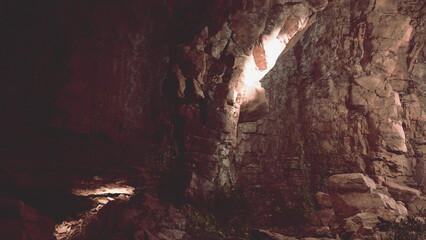A cavernous space is illuminated by an otherworldly glow, highlighting the rugged stone walls and the natural greenery below. Shadows dance in the tranquil atmosphere, enhancing the enchanting scene.