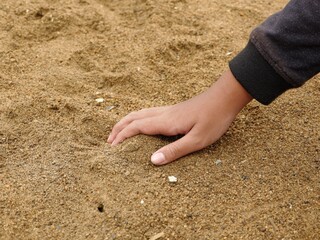 Detail shot of textured sand surface with tiny pebbles and a person's hand in a dark sleeve, emphasizing touch, play, and natural materials.