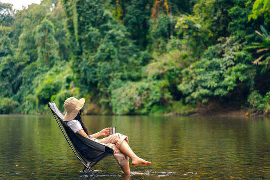 A woman sits on a camp chair on the river. She held a stainless steel glass and looked at the view in the morning. The water is clear and pure with flow. The background is a forest with lush trees.