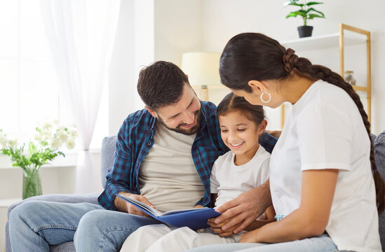 Happy family reading book together at cozy home. Happy father, mother and daughter sit on sofa to read storybook, explore pictures and enjoy fun time and relaxation in living room at weekend - Powered by Adobe
