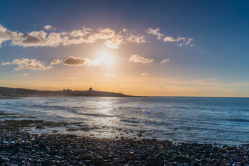 Fototapeta premium Summer sunset in Shimen District, New Taipei City, Taiwan, with golden sunlight reflecting on calm sea, rocky shore, and distant hill silhouette.
