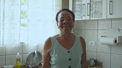Smiling Elderly Woman Standing In A Home Kitchen Looking At The Camera With A Joyful Expression, White Cabinets And Tiled Walls In The Background