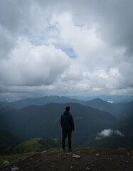 A lone figure stands on a rocky hill, overlooking misty mountains under a dramatic sky filled with clouds.
