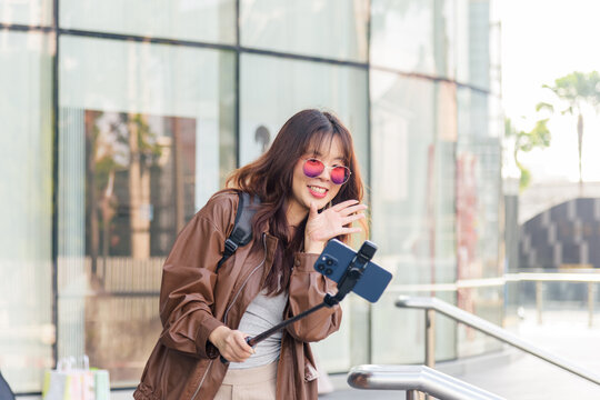Asian woman, fashion lifestyle blogger, smiles bright, vlogging outside shopping mall, creating social media content, wearing stylish brown jacket and trendy sunglasses in urban city.