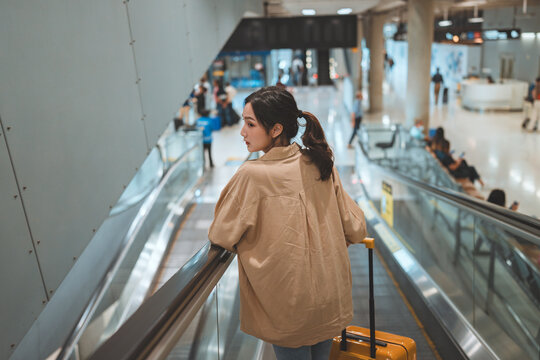 Rear view Asian tourist woman traveller with simple suitcase stand on moving walkway in airport terminal, Tourist journey trip concept