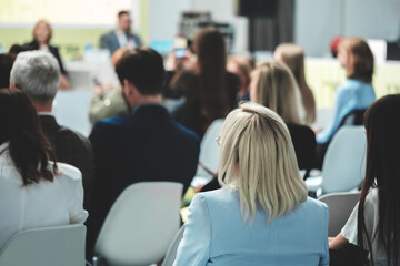 A business woman with blond hair in a blue suit sits at a business presentation in a conference room and listens to the speaker.