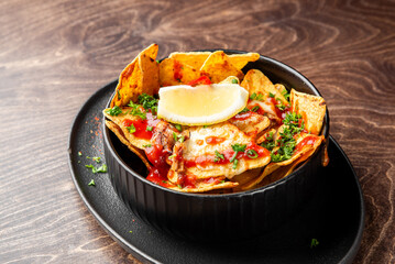 Delicious loaded nachos served in a black bowl on a dark wooden table. Topped with melted cheese, meat, red sauce, fresh parsley, and a lemon wedge. Perfect as a snack or appetizer.