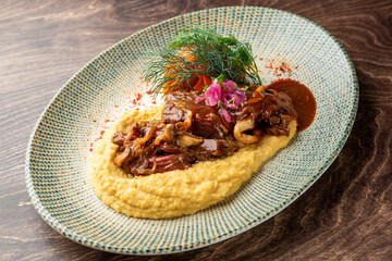 A gourmet dish of slow-cooked beef cheeks and mushrooms served over creamy polenta or corn puree, garnished with pickled onions and dill on a rustic plate