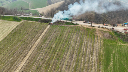 Aerial View of Burning Grapevine Branches and Brushwood Smoldering Amidst the Rolling Piacenza Hills Agricultural Landscape in Northern Italy Under a Clear Sky