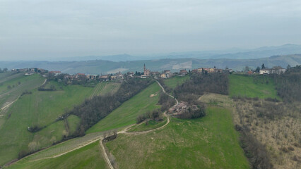 Obraz premium Panoramic vista of the rolling Piacenza hills in Italy revealing clusters of rural architecture and verdant landscapes under a hazy sky with distant mountain ranges and a small village nestled on the