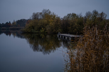 Jesienny krajobraz z pomostem nad jeziorem – spokojna tafla wody i odbicia drzew w pochmurny dzień © Kacper