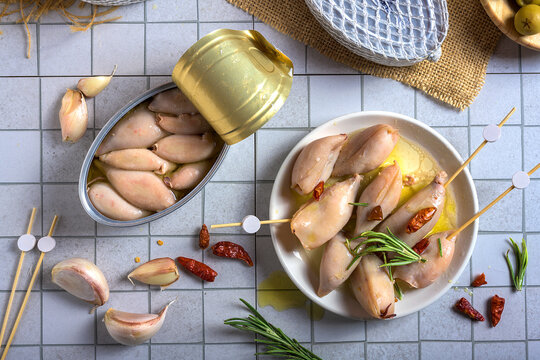 Top view of gourmet canned squid in oil served as Spanish tapas. Arranged on a bright tile surface with garlic, rosemary, and chili peppers. Ideal for Mediterranean cuisine and seafood concepts