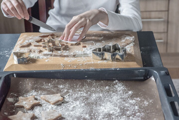 Hands actively cutting and preparing gingerbread cookie dough with metallic cutters and a knife on a wooden board. Traditional, focused, cozy holiday baking process.