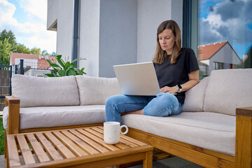 Woman sitting on terrace sofa using laptop with coffee mug on wooden table. Female freelancer working remotely from home