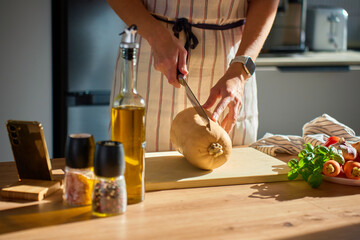 Person in apron cutting butternut squash on wooden board in kitchen. Close-up of woman slicing fresh pumpkin with fresh vegetables and herbs on table. Concept of healthy eating and home cooking