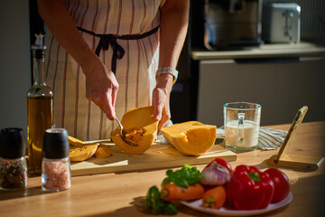 Woman scooping pumpkin seeds and using smartphone in modern kitchen with sunlight. Person in apron preparing butternut squash for roasting. Concept of home cooking and online recipe