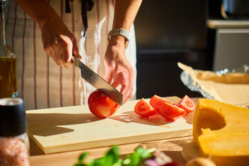 Close-up of woman cutting tomato on wooden board in kitchen with vegetables and pumpkin on table. Concept of organic food and healthy cooking