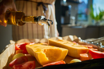 Woman in apron preparing pumpkin and vegetables on baking tray pouring olive oil before roasting. Female cooking healthy vegetarian meal in home kitchen, making pumpkin soup