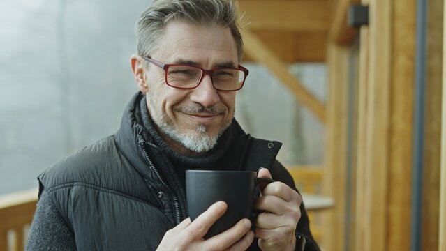 Happy mature man in sweater and glasses smiling while holding warm mug on cozy porch, enjoying a winter or autumn day. Mid adult middle aged male in 50s drinking hot tea or coffee outdoor.