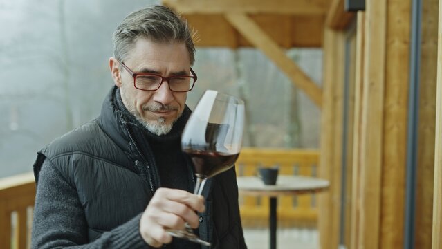Senior winemaker enjoying aroma of red wine in glass, standing at wooden balcony of winery. Mature man drinking, tasting and analyzing quality of wine.