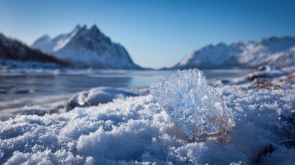 Obraz premium An ice crystal rests on soft snow beside a calm lake. Majestic mountains rise in the distance under a bright blue sky. The scene captures the beauty of winter tranquility.