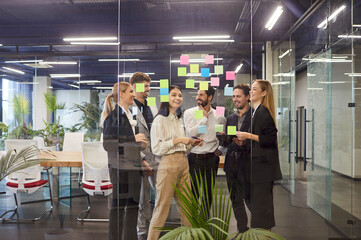 Group of business professionals collaborating in modern office, brainstorming ideas with sticky notes on glass wall. Smiling team of colleagues discussing strategies, showing teamwork and creativity.
