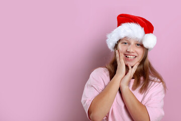 Excited pretty 11-year-old girl in pink T-shirt and Santa hat posing on pink background