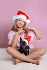 Cute holiday photo — little girl in pink making a heart shape, while her Chihuahua looks dramatically evil. Funny Christmas portrait of a girl next to a grumpy Chihuahua in Santa hats 