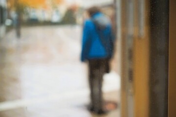 Abstraktes Motiv mit Rückenansicht von Frau mit blauem Anorak, grauer Schultertasche und grauer Jeanshose steht in Fußgängerzone in Stadt bei Regen am Nachmittag im Herbst