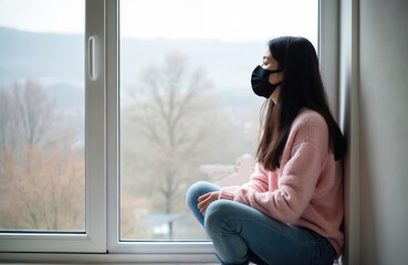 Sad Asian woman wearing black face mask sits alone at home by window. She looks outside feeling lonely stressed during pandemic quarantine. Mental health issues and isolation are evident.