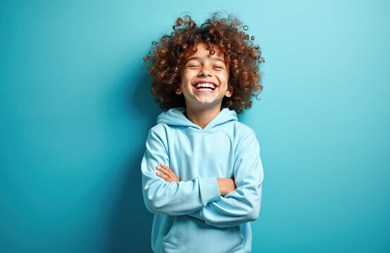 Young boy with curly brown hair smiles widely with arms crossed. Wears light blue hoodie against plain blue backdrop. Child looks joyful, confident, possibly reflecting happiness success in school