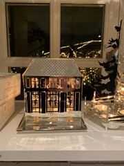 Metal Candle Holder House with Warm Candlelight Glow at Night; metal candle holder in the shape of a house illuminated by warm candlelight, creating cozy reflections on a window at night