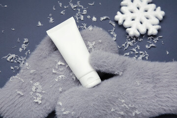 Winter skincare concept with grey mitten and moisturizing cream, surrounded by soft snow on blue.