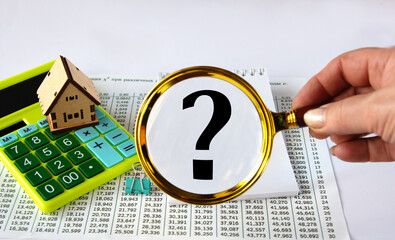 Question mark on a white sheet of paper with a calculator and a wooden house in the background