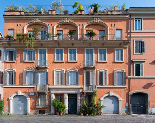 A warm orange façade apartment building lines a cobbled street with shuttered windows, decorative balconies, lush plants and rooftop greenery, Rome, Italy