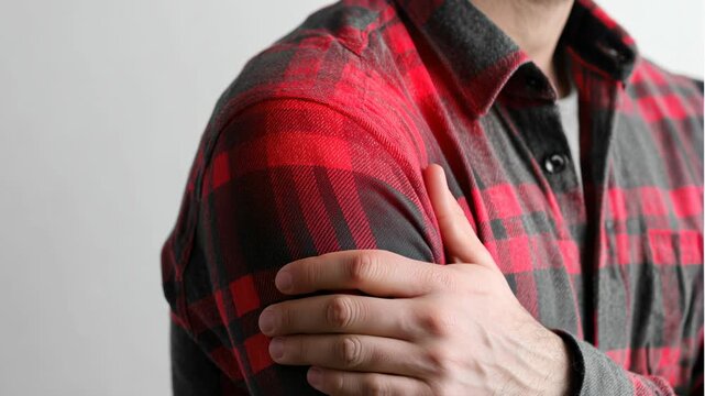 A young  man with dark hair wearing a red and gray plaid shirt holds his shoulder, indicating discomfort or pain.
