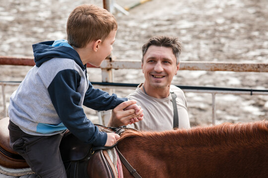 Happy father walking next to his young son riding a brown horse on a bridle path in an equestrian training center on an autumn day