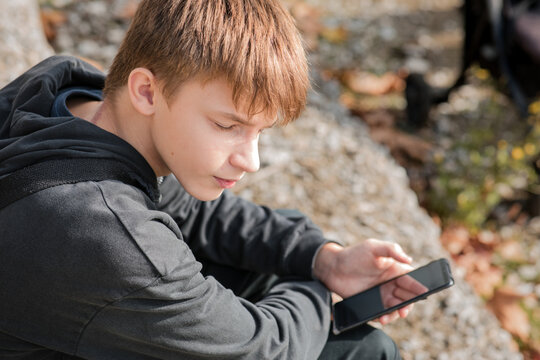 Young teenage boy sitting outdoors, focused on using a black smartphone with a blank screen on a sunny day